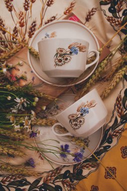 Sarı masa örtüsünün üzerinde iki tane antika bardak var. Bardakların yanında bir buket kır çiçeği var..