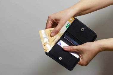 Avro banknotları alırken bir kişi açık siyah bir cüzdan tutuyor. İçeride, kartlar ve düzenli bölmeler görülebilir durumda. Banknotlar 50 avro gibi görünüyor ve kısmen kaldırılmış durumda. Bu da ödeme veya nakit çekilmesi anlamına geliyor..