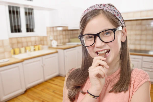 Schattig preteen meisje met bril eet chocolade, keuken backg — Stockfoto