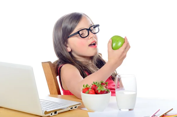 En liten flicka sitter vid sitt skrivbord äta ett äpple — Stockfoto