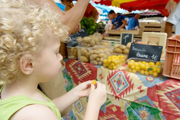 A little blond boy eats plums on the market — Stock Photo, Image