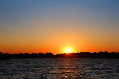 Güneşli arka planda, gökyüzünün ve şehrin saf renklerinden oluşan parlak arkaplanlı deniz üzerinde harika bir günbatımı diski.