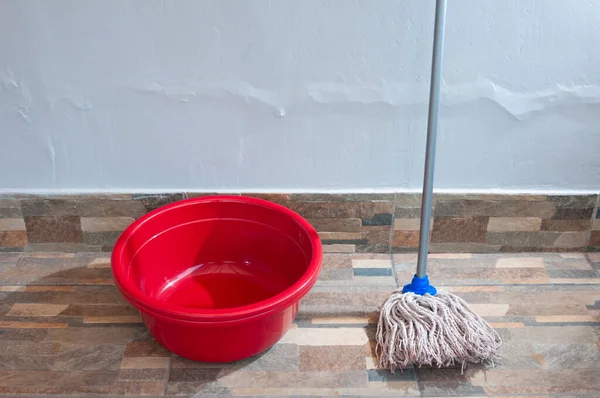 Metal saplı bir paspasın yanında parlak kırmızı plastik bir kova ve gri iplik lifleri desenli fayans zemininde gri bir duvara dayanıyor..