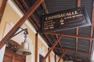 Chimbacalle tren istasyonundaki ahşap bir yapının tavanından sarkan, bronz bir çanın yanında asılı duran, siyah ahşap bir tabelanın düşük açılı görüntüsü.