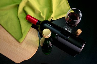 Kırmızı şarap şişesi, dolu bir bardak, küçük bir şişe, bir mantar ve yansıtıcı siyah bir yüzey üzerinde yeşil bir masa örtüsü bulunan kuş bakışı bir manzaradan hareketsiz bir hayat..