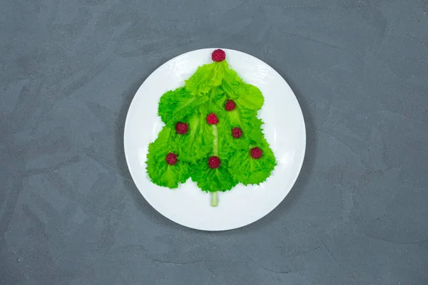 Choinka wyłożona jest zielonymi liśćmi sałaty, ozdobionymi czerwonymi malinami na białym talerzu. Jedzenie na Nowy Rok. Dekoracja stołu. Widok z góry. — Zdjęcie stockowe