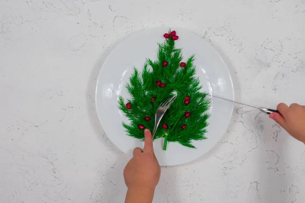 A árvore é forrada com folhas de endro verde, decoradas com bagas de romã vermelha em um prato branco. As mãos das crianças estão segurando um garfo e uma faca. Comida para o Ano Novo. — Fotografia de Stock