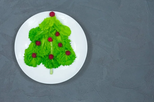 Choinka wyłożona jest zielonymi liśćmi sałaty, ozdobionymi czerwonymi malinami na białym talerzu. Jedzenie na Nowy Rok. Dekoracja stołu. Widok z góry. Miejsce napisania. — Zdjęcie stockowe