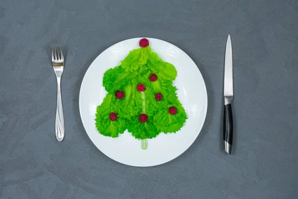 Choinka wyłożona jest zielonymi liśćmi sałaty, ozdobionymi czerwonymi malinami na białym talerzu widelcem i nożem. Jedzenie na Nowy Rok. Dekoracja stołu. Widok z góry. — Zdjęcie stockowe