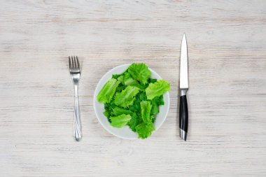 Tahta bir masada çatal ve bıçakla beyaz bir kasede yeşil salata. Düzgün beslenme, diyet.