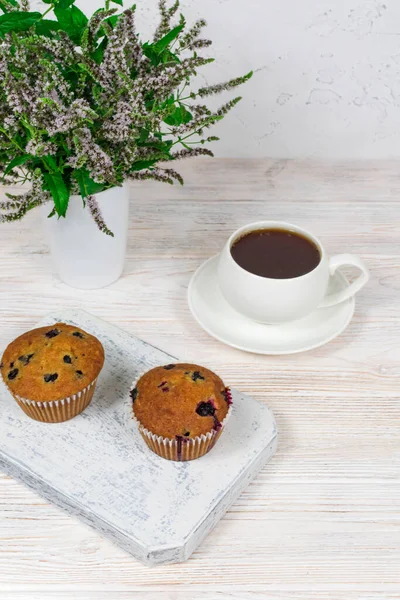 Cupcakes com groselhas pretas em uma placa de corte de madeira branca contra o fundo de uma xícara de chá. — Fotografia de Stock