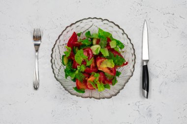 Green vegan salad made from a mixture of green leaves and tomatoes in a salad bowl with, a fork and knife on a white stone table.