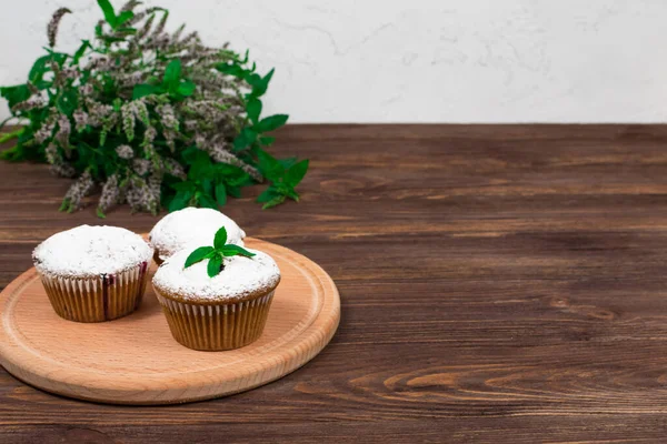 Blackcurrant cupcakes with a white cup of tea on a background of flowers and mint leaves, on a round board on a wooden background. — Stock Photo, Image