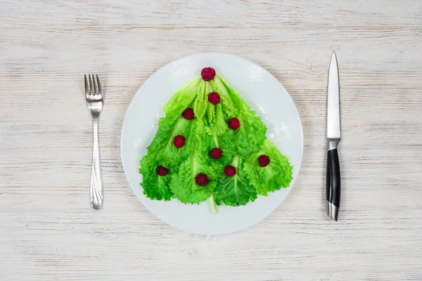 A árvore de Natal é forrada com folhas de alface verde, decoradas com framboesas vermelhas em uma placa branca com um garfo e faca. Comida para o Ano Novo. Decoração de mesa. Vista de cima. — Fotografia de Stock