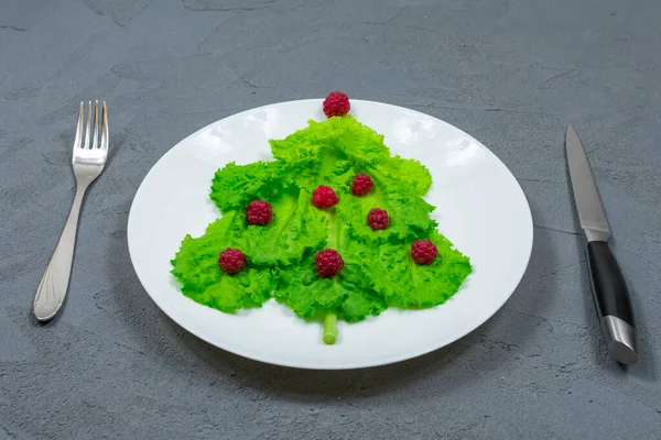 A árvore de Natal é forrada com folhas de alface verde, decoradas com framboesas vermelhas em uma placa branca com um garfo e faca. Comida para o Ano Novo. Decoração de mesa. Vista de cima. — Fotografia de Stock