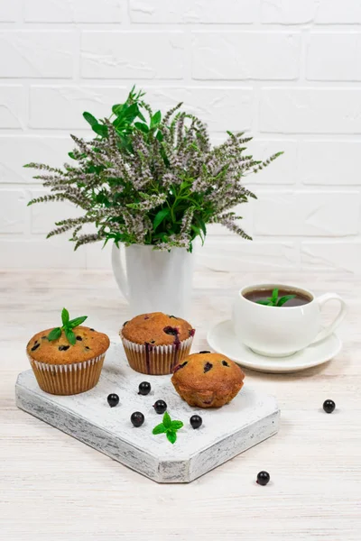 Cupcakes com groselhas pretas em uma placa de corte de madeira branca contra o fundo de uma xícara e flores de hortelã. — Fotografia de Stock