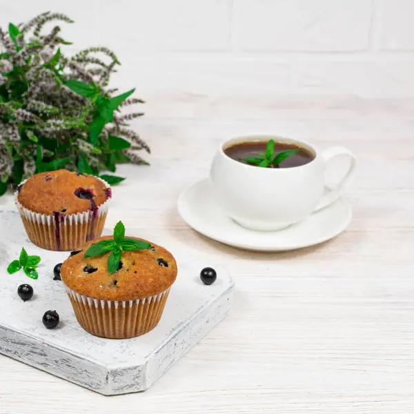 Cupcakes com groselhas pretas em uma placa de corte de madeira branca contra o fundo de uma xícara e flores de hortelã. — Fotografia de Stock