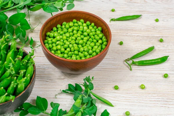 Fresh green peas in a bowl against the background of leaf shoots, sprigs of young green peas on a white wooden table. View from above. Proper nutrition, vitamins. Close-up. Selective focus. Defocus.