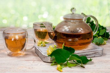 Cam çaydanlık ve iki bardak çay. Üzerinde ıhlamur ağacı olan ahşap bir masa, bulanık bir zemin ve ıhlamur yaprakları, çiçekler. Çay demliyorum..