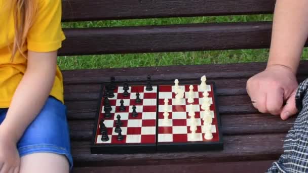 Petite fille joue aux échecs avec papa dans le parc sur un banc à l'air frais. Le jour des échecs. Jeux éducatifs, éducatifs en plein air. Développement de l'enfant. — Video