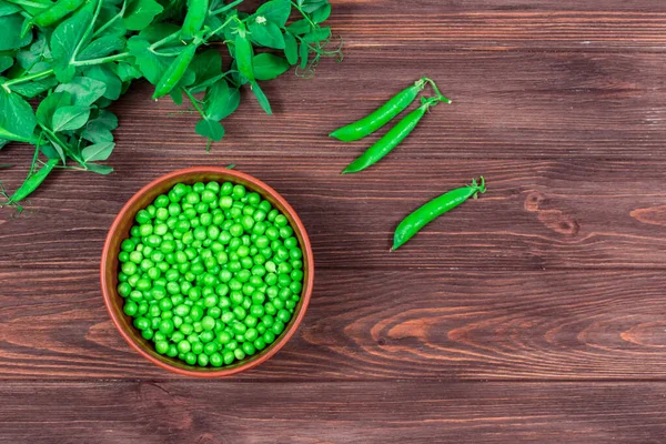 Guisantes verdes frescos en un tazón contra el fondo de los brotes de hojas, ramitas de guisantes verdes jóvenes en una mesa de madera oscura. Vista desde arriba. —  Fotos de Stock