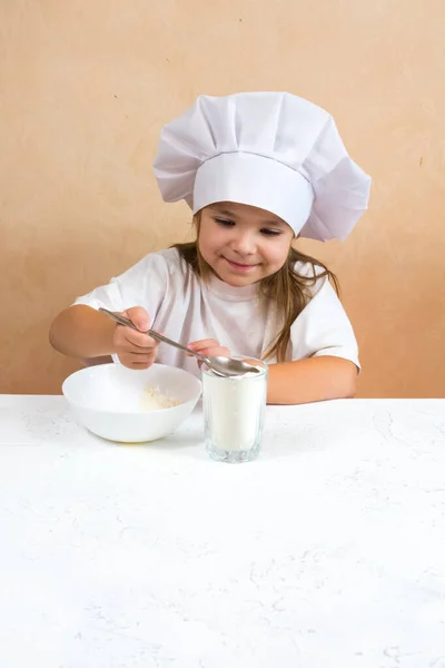 Una niña vestida de cocinera amasa la masa. Cocinar concepto de estilo de vida infantil. El niño ama, se divierte, estudia y juega en la cocina — Foto de Stock