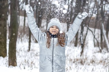 Kışlık ceket giymiş ve örgü şapka takmış gülümseyen bir kız, karlı orman eğlencesinin tadını çıkarır..