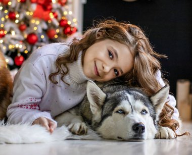 A young girl rests with a husky by a decorated Christmas tree, warm, intimate holiday scene at home.