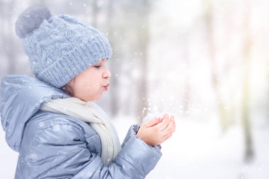 Mavi kışlık paltolu ve eşarplı bir çocuk huzurlu, karlı bir parkta kar üflüyor..