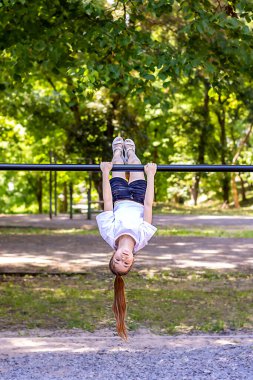 Genç bir kız, yapraklı bir parkta, yatay bir barda baş aşağı asılı, gülümsüyor ve açık hava eğlencesine eğleniyor..