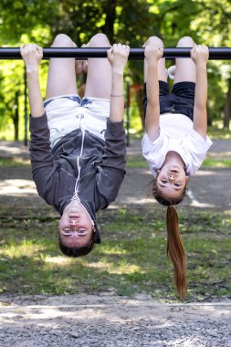 İki kız oyun parkındaki bir barda baş aşağı asılı, yazın dışarıda gülüp oynuyorlar..