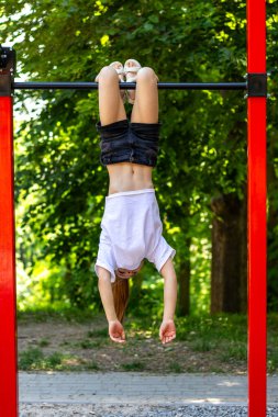 Genç bir kız yeşil ağaçların arasında oyun parkındaki bir barda baş aşağı asılı, neşeli yaz eğlencesinin tadını çıkarıyor..