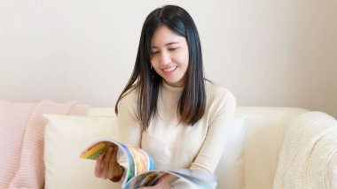 Genç Asyalı kadın evdeki kanepede kitap okuyor.