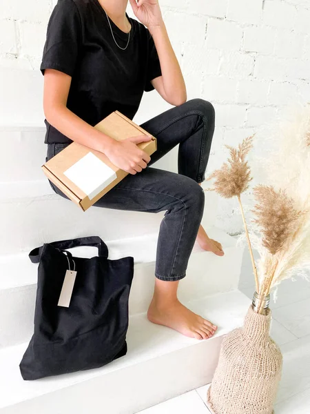 Hermosa mujer joven en camiseta negra y jeans con bolsa de mano y caja de cartón — Foto de Stock