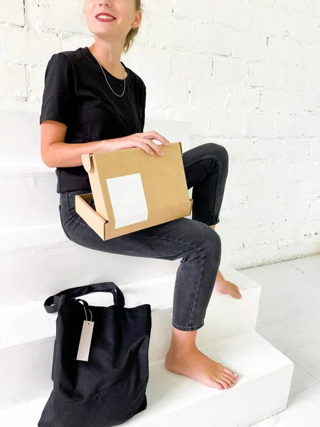 Hermosa mujer joven en camiseta negra y jeans con bolsa de mano y caja de cartón — Foto de Stock