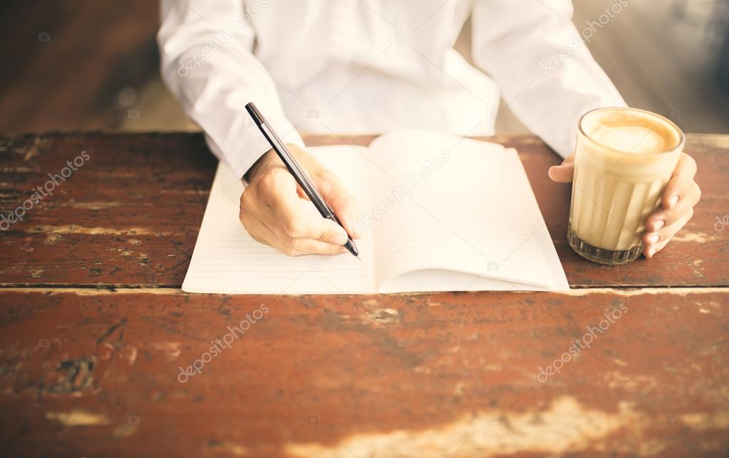 Man writing in notepad with cup of coffee in left hand on wooden