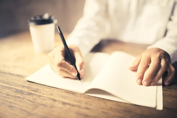 Κορίτσι γράφει στο Σημειωματάριο (Notepad) σε ένα ξύλινο γραφείο με ένα φλιτζάνι καφέ — Φωτογραφία Αρχείου