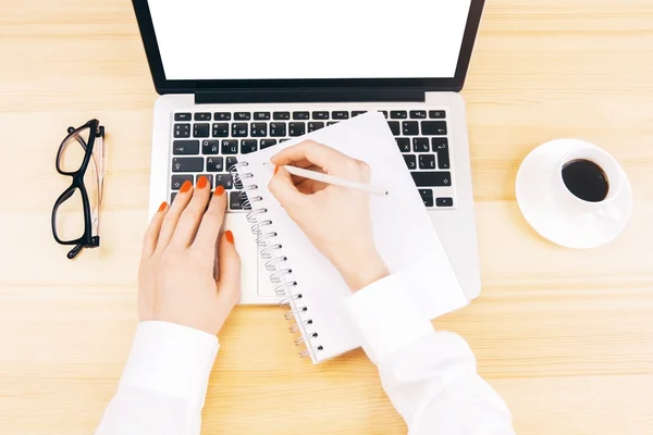 Vedere de sus a mâinilor de sex feminin scris în notepad pe birou din lemn cu laptop alb gol, ochelari și ceașcă de cafea. Mock up — Fotografie, imagine de stoc