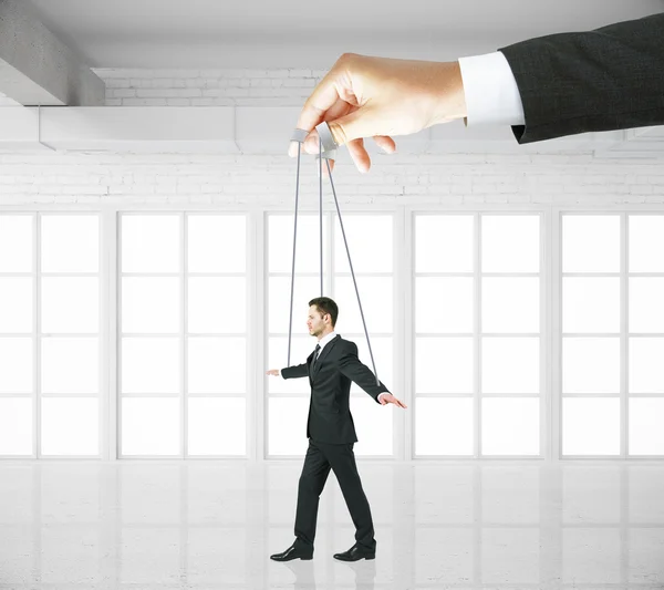 Hand manipulating businessman puppet on ropes in white brick interior. Concept of control — Stock Photo, Image