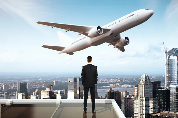 Travel concept with businessman on roof looking at flying by airplane. City and sky background — Stock Photo, Image