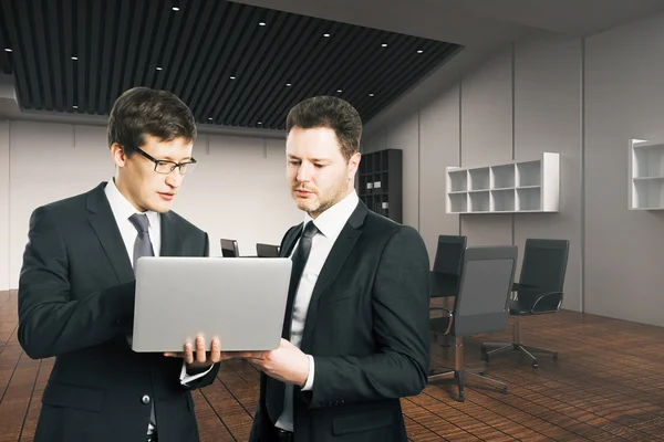 两个年轻的商人与笔记本电脑站在现代化的办公室内部。团队合作理念 — 图库照片
