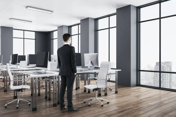 Businessman standing in luxury office interior with panoramic city view,table with computers and big window. Coworking workplace concept