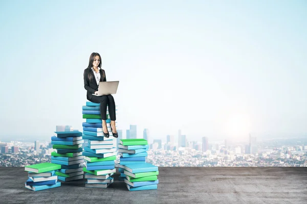 Young businesswoman with laptop computer sitting on books on city background. Growth, knowledge and vision concept