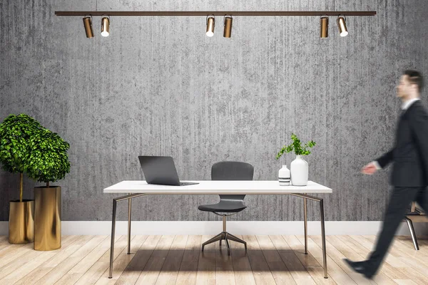 Businessman walking in director\'s office with computer on table and blank gray wall. Workplace and worker concept.