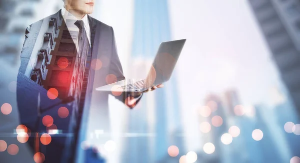 Remote working concept with employer in black suit with opened laptop at blurry city skyscrapers background. Double exposure