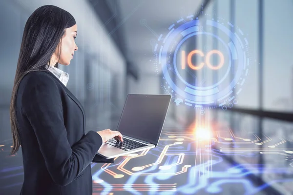 Businesswoman with a tablet touches a hologram with ico letters in an office corridor, trade and market concept