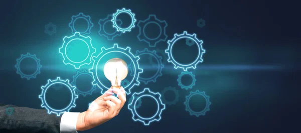 Start up and idea concept with digital gears and glowing light bulb in man hand on abstract dark background