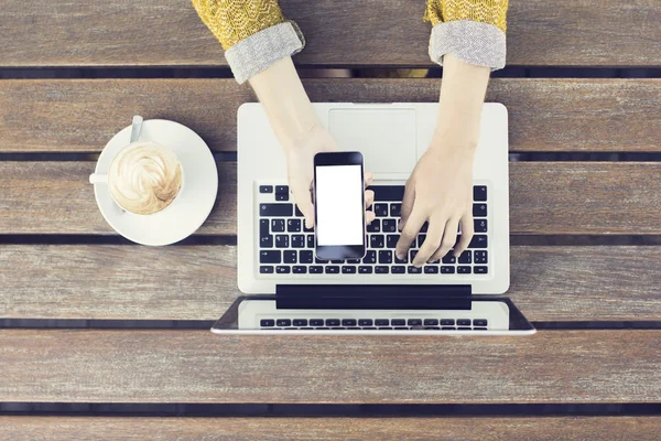 Menina com smartphone em branco, laptop e cappuccino na mesa de madeira — Fotografia de Stock