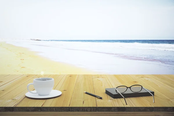 Kopp kaffe notebook och glasögon på ett träbord på stranden — Stockfoto