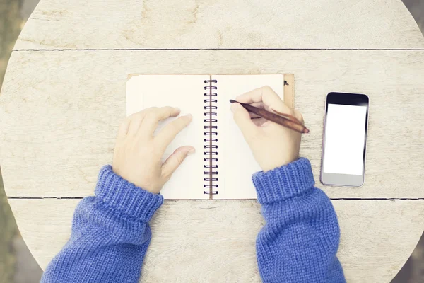 Meisje schrijft in notitieblok aan een houten tafel en een lege cel-telefoon — Stockfoto